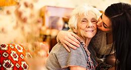 An older woman with short white hair smiles as a young woman with long dark hair leans over to give her a kiss on the cheek.